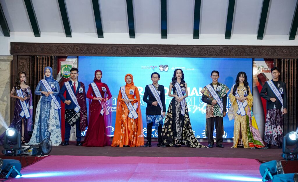 Group of pageant contestants in evening gowns and suits wearing sashes and numbered badges on a stage with a blue backdrop
